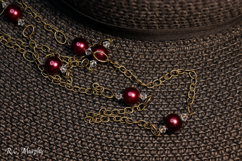 A 31 inch bronze chain in a closed loop sprinkled with 7 burgundy faux pearls framed by crystals. The necklace sits on the brim of a black sunhat.