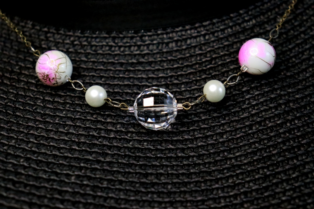 A disco ball shaped crystal beads is framed by small glass pearls and larger white glass beads with pink and metallic paint splatter. Antiqued brass colored chain. The necklace rests on a black sunhat brim.